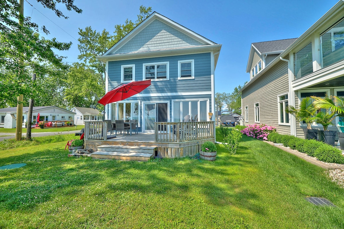 The exterior of the cottage features a light blue facade with a spacious deck equipped with a red umbrella. Lush green grass surrounds the deck, while nearby colorful flowerbeds add vibrancy. The structure is two stories high, set within a peaceful neighborhood.