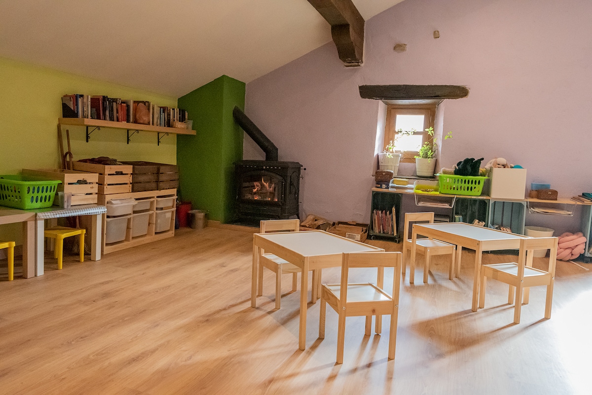 A brightly colored room features light wooden flooring and a cozy fireplace. Small tables and chairs are arranged for various activities. Shelving holds books and storage baskets, while a window allows natural light to fill the space, complemented by a potted plant.