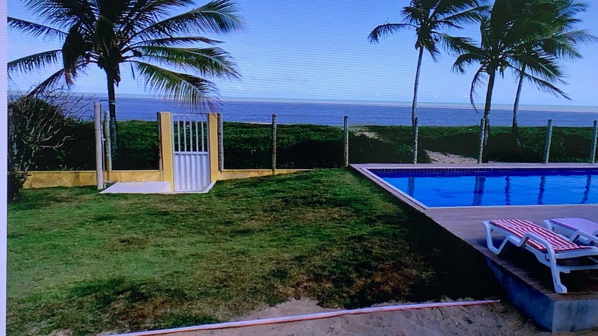 A serene outdoor area features a clear swimming pool surrounded by a wooden deck. Lush green grass extends towards a fence near the ocean. Palm trees sway gently in the breeze, and a gate offers access to the beach.