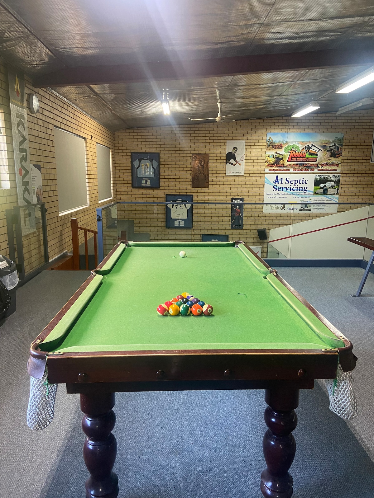 A well-maintained billiards table is positioned at the center of the room, featuring a bright green felt surface and a collection of colorful pool balls arranged at one end. Soft lighting highlights the space, with artwork and signage accentuating the brick wall background.