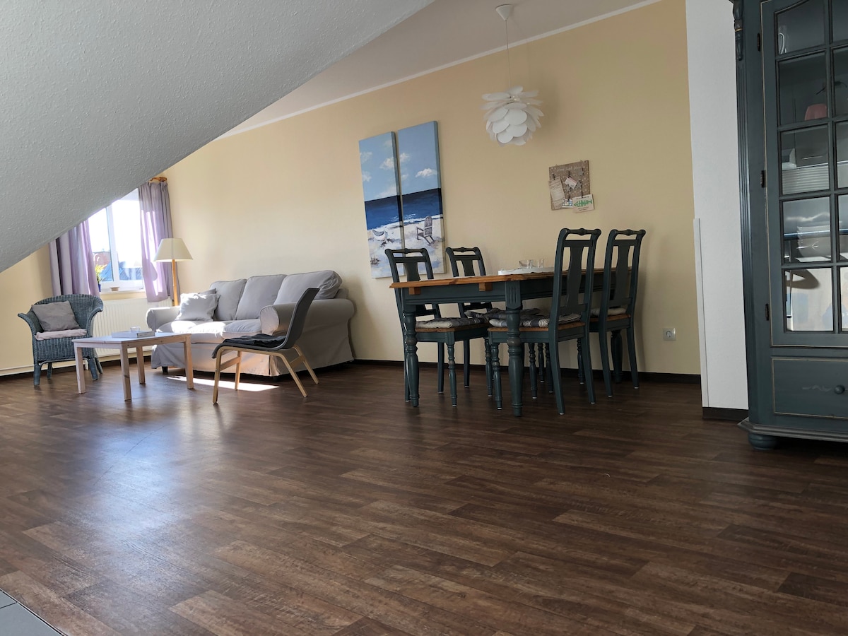 A bright and airy living space is seen featuring a comfortable sofa and a dining table with six chairs. Natural light is allowed to flow in through large windows, enhancing the warm tones of the walls and flooring.