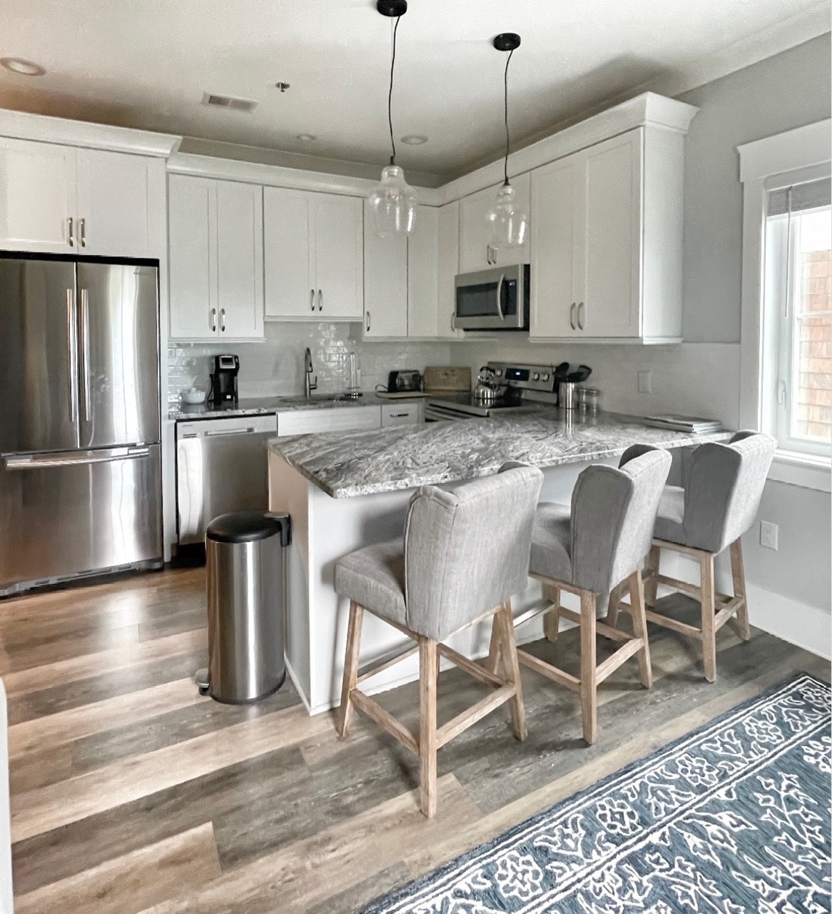 A fully equipped kitchen features modern, white cabinetry and a spacious granite countertop. Three upholstered bar stools are positioned at the island. Stainless steel appliances, including a refrigerator and microwave, complement the room. Natural light streams in through the adjacent window.