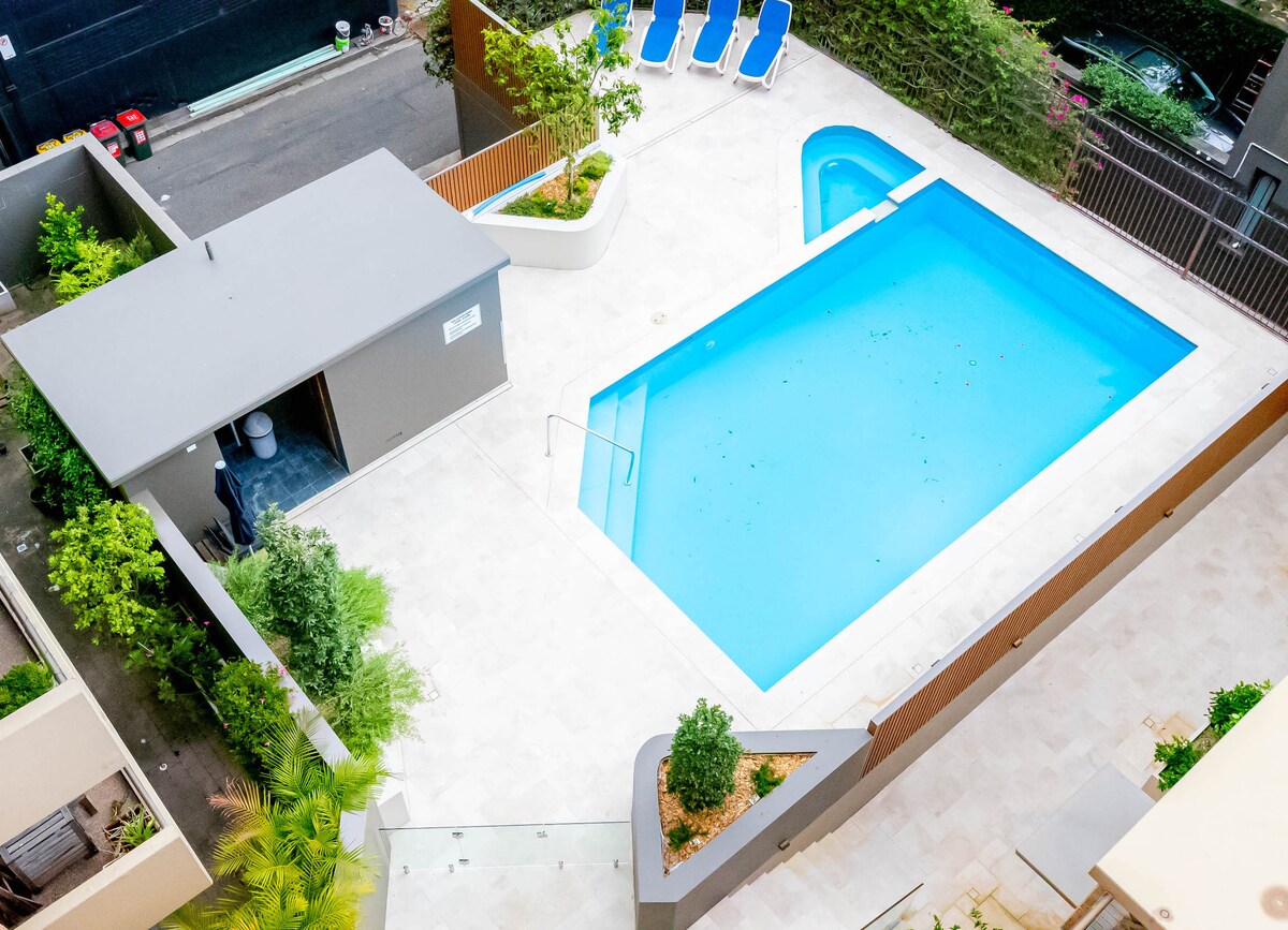 A modern outdoor pool area is depicted, featuring a rectangular pool surrounded by a tiled deck. Sun loungers line the side of the pool, while landscaped greenery adds a touch of nature. A small poolside shelter houses changing facilities.