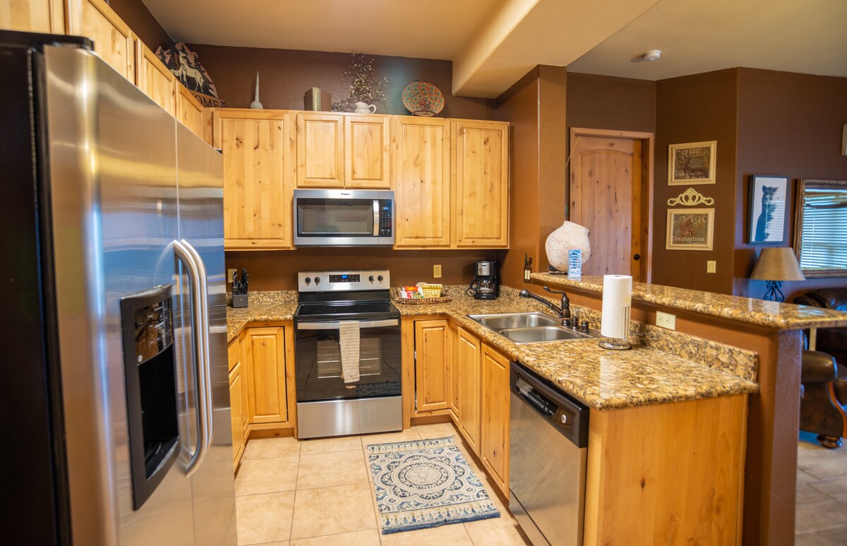 A fully equipped kitchen features wooden cabinetry and granite countertops. Stainless steel appliances, including a refrigerator, oven, and microwave, are visible. The kitchen island is complemented by a decorative rug, and ample natural light is provided through a nearby window.