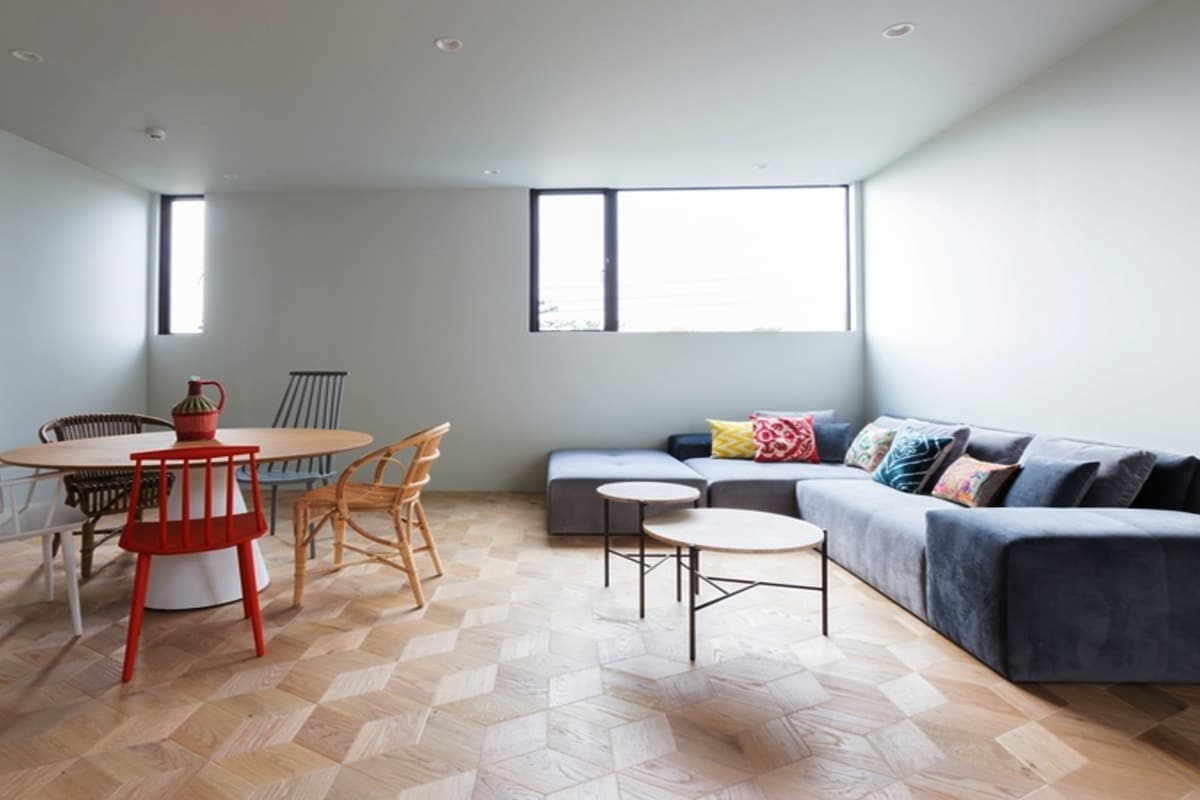 A spacious living area features a mix of seating options, including a large, modern sofa adorned with colorful cushions. A round dining table sits nearby, surrounded by various chairs. Natural light floods in through large windows, illuminating the hexagon-patterned wooden floor.