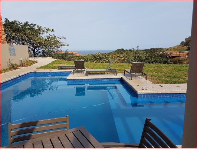 A tranquil pool area is shown, featuring a rectangular pool with crystal-clear water. Lounge chairs are positioned around the pool, offering views of the ocean and surrounding landscaped gardens. The outdoor patio space is well-maintained, promoting relaxation in a serene environment.