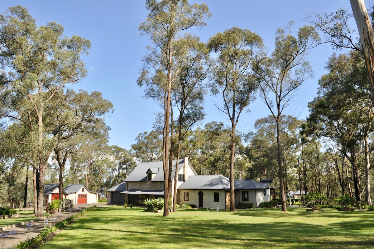 A spacious bush retreat is surrounded by tall trees and a well-maintained lawn, providing a serene setting. The architecture features a combination of light and dark elements, with multiple rooflines and a welcoming façade visible under a clear blue sky.