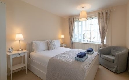 A bright and airy bedroom features a spacious bed adorned with neatly arranged linens. Two bedside tables with lamps are positioned on either side. A cozy armchair sits in the corner, and large windows provide natural light, framed by light curtains.