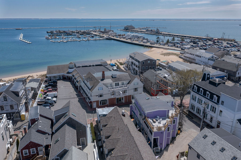 An aerial view captures the vibrant harbor and sandy beach alongside the coastal town. Various boats are docked at the marina, while the buildings of the town showcase a mix of residential and commercial spaces, all under a clear blue sky.