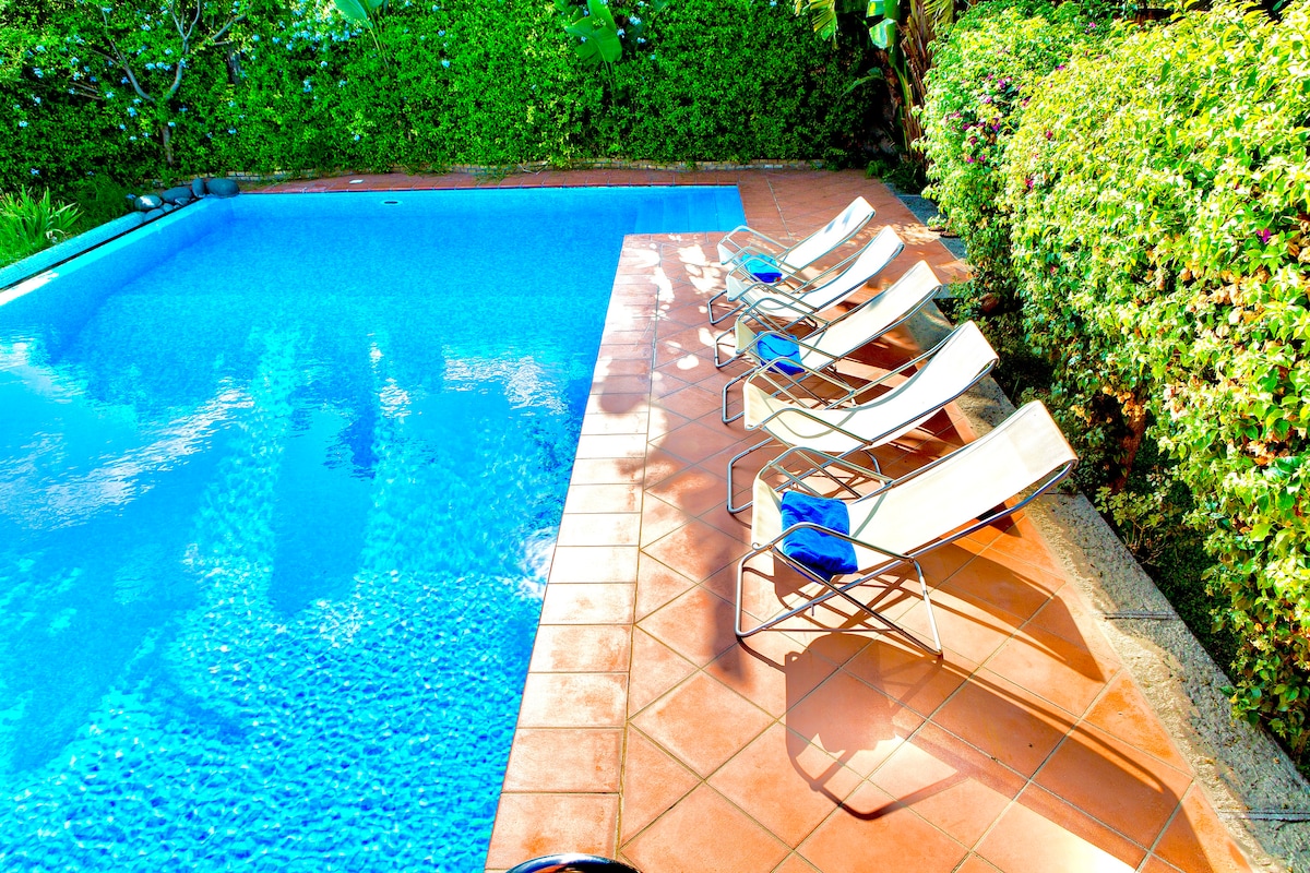 A clear blue pool is surrounded by lush greenery, offering a relaxing atmosphere. Several lounge chairs are positioned along the edge, inviting guests to enjoy the sun. The tiled area surrounding the pool provides ample space for leisure and relaxation.