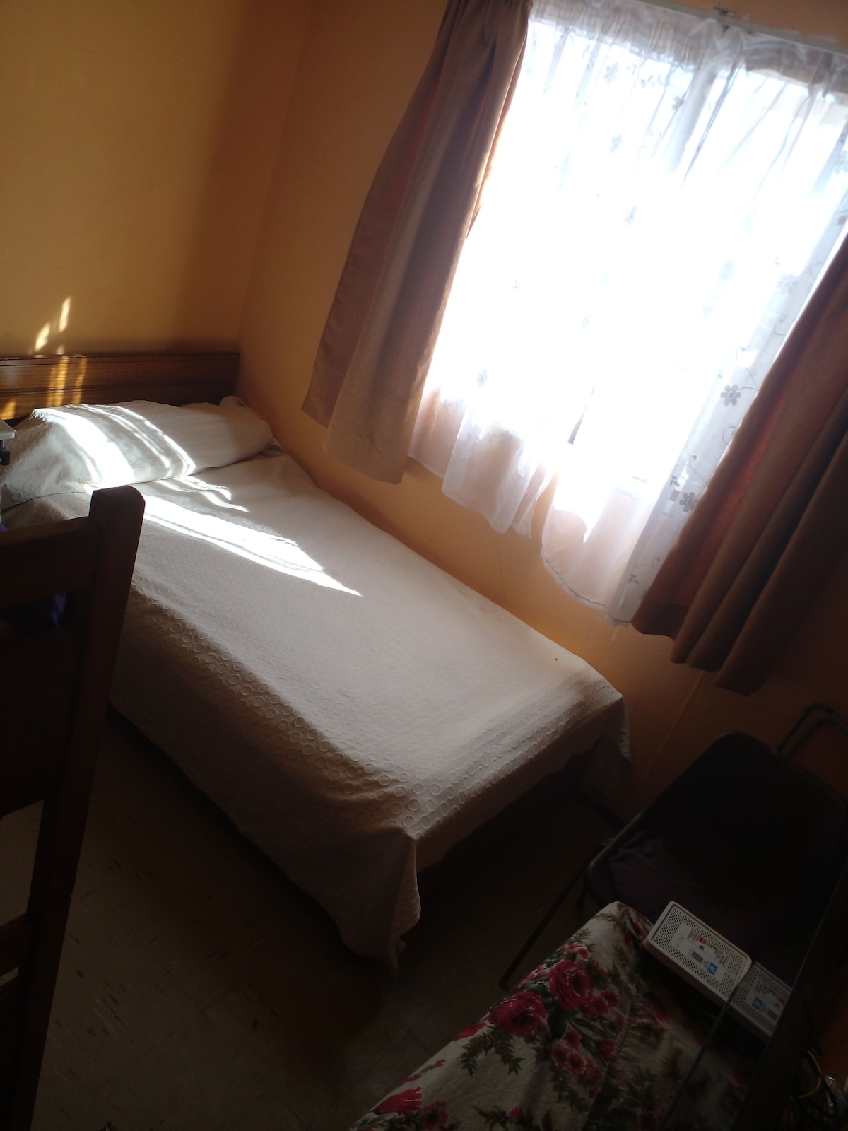 A cozy sleeping area features a single bed covered with a light blanket. Soft natural light fills the room through a window adorned with sheer curtains. Warm-toned walls contribute to a calm atmosphere, and a chair is positioned nearby.
