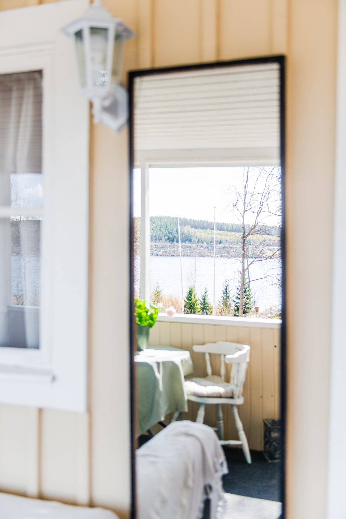 A reflection in a tall mirror reveals a cozy terrace overlooking the water. A small table is set with a green tablecloth, accompanied by light-colored chairs. The distant landscape features trees lining the shore, while the calm water adds a serene backdrop.