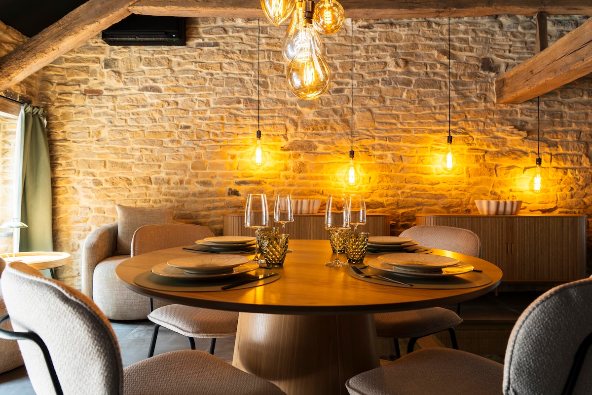 A dining area is showcased, featuring a round wooden table surrounded by light-colored chairs. Warm stone walls are illuminated by pendant lights with exposed bulbs. Elegant table settings with glassware and decorative elements enhance the inviting space.