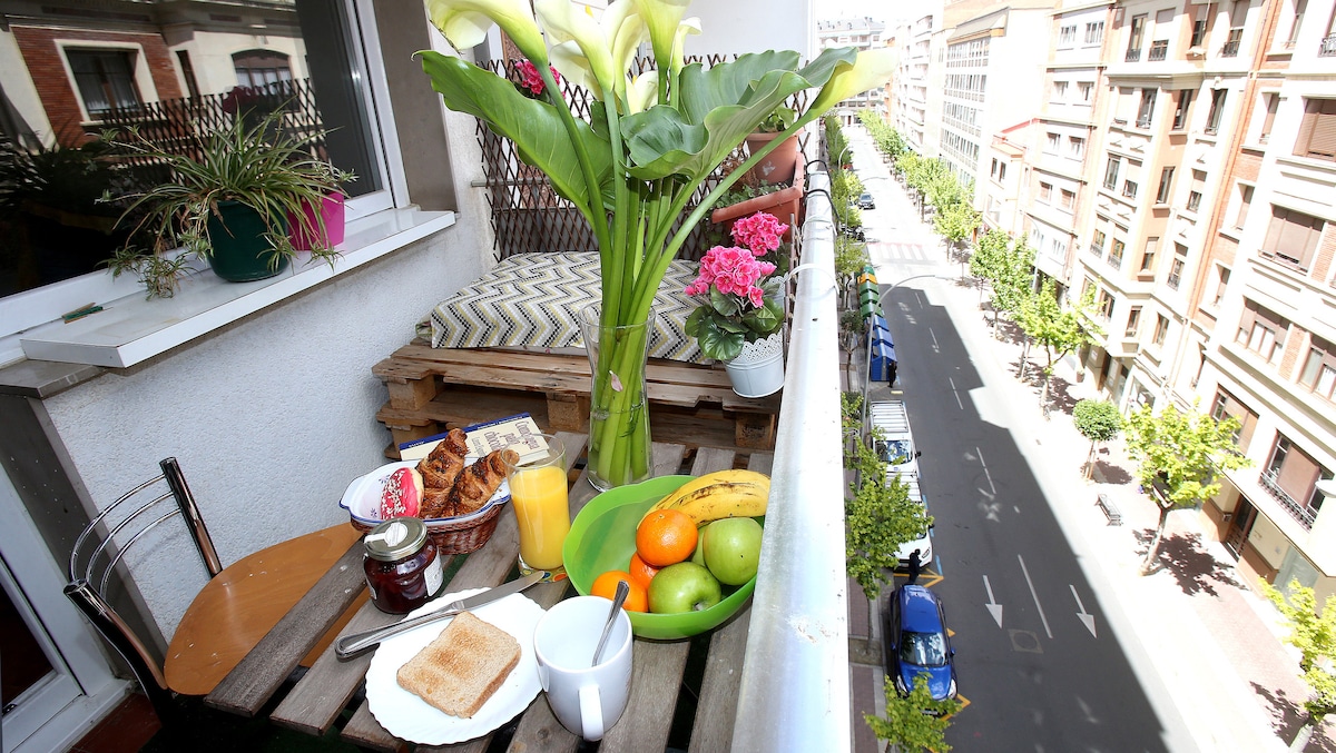 A private balcony is depicted with a wooden table set for breakfast. Fresh fruit, pastries, and beverages are arranged alongside a bouquet of flowers. The view includes tree-lined streets and nearby buildings, suggesting a vibrant urban atmosphere.
