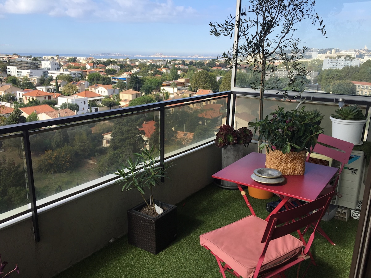 A balcony area features a small pink table with two matching chairs, surrounded by potted plants. The view includes distant greenery and rooftops, with the sea visible beyond. The flooring appears to be a grass-like material, enhancing the inviting atmosphere.