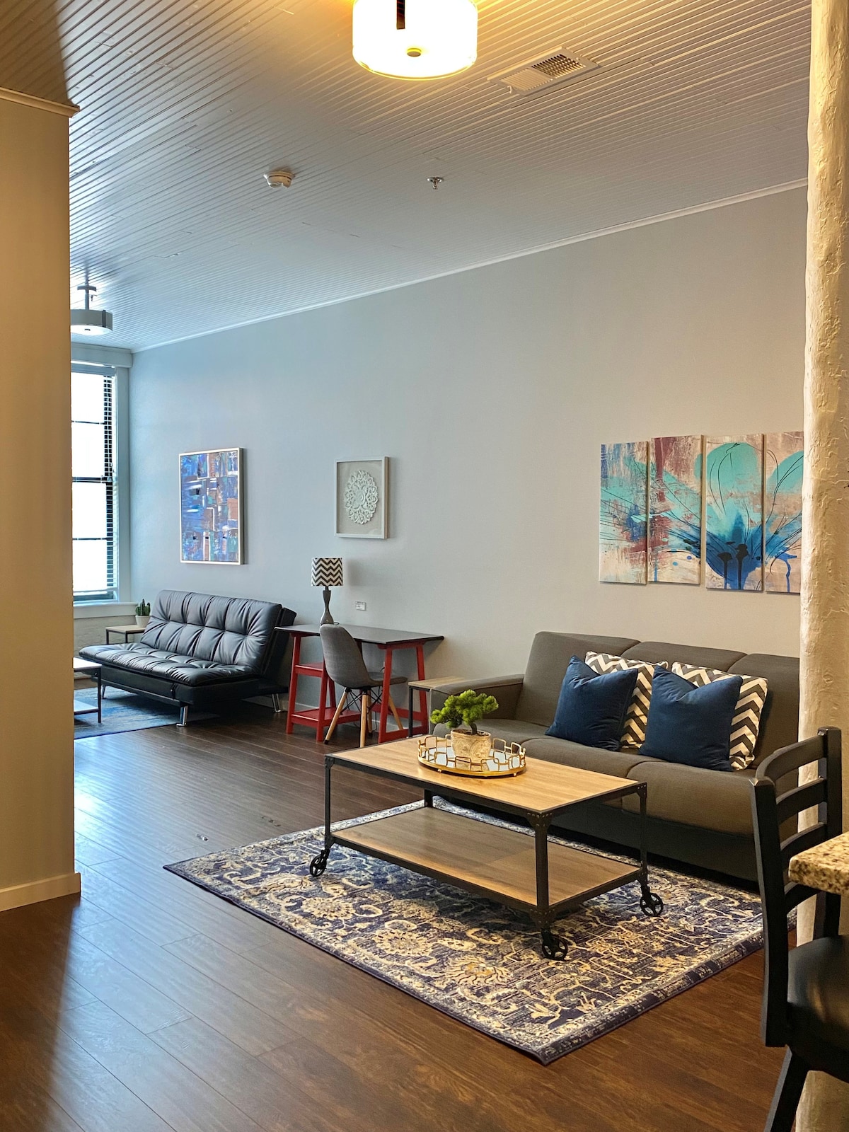 The living area features a modern futon and a coffee table with decorative trays. Artwork adorns the walls, complemented by a small lamp and a potted plant. A rug enhances the hardwood flooring, while large windows allow for ample natural light.