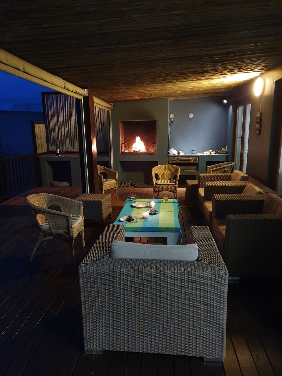 An outdoor seating area features comfortable wicker furniture arranged around a soft blue tablecloth. A cozy fireplace provides warmth, with soft lighting accentuating the inviting space. Wooden decking adds a natural touch, creating an ideal spot for relaxation in the evening.