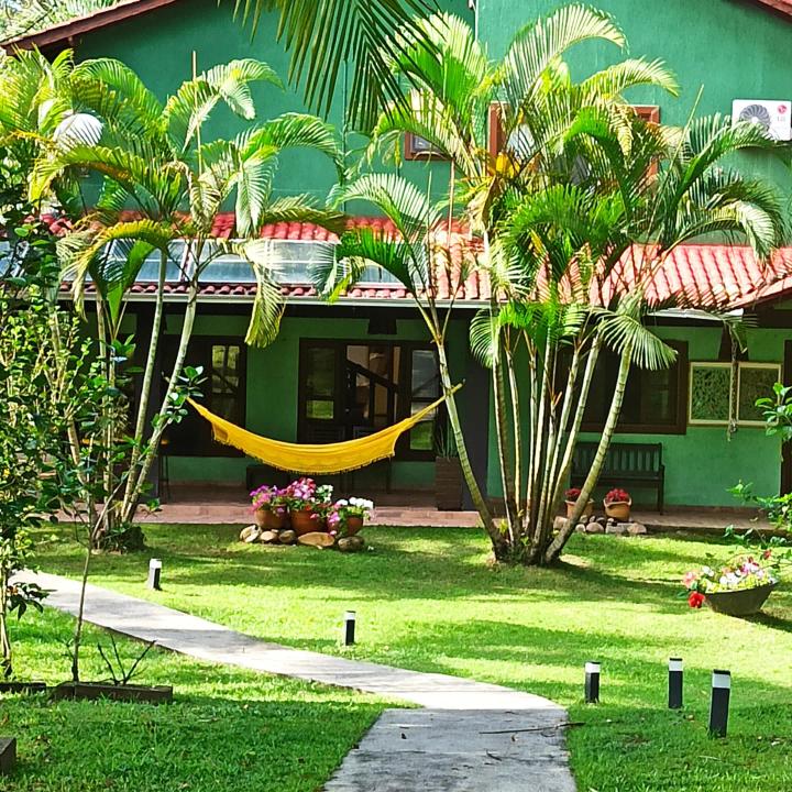 A vibrant green pousada is surrounded by palm trees and lush gardens. A pathway leads to the entrance, while a hammock is gently strung between the trees. Flower pots add color to the outdoor space, creating a tranquil environment.