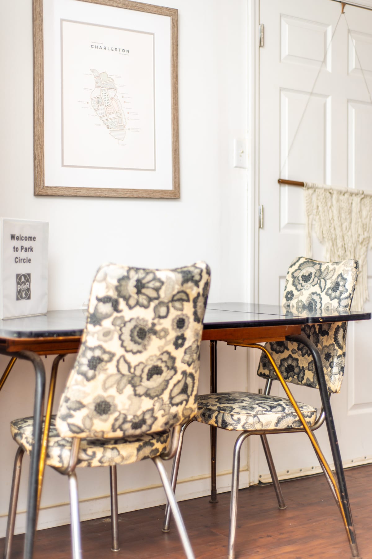 A charming dining area is presented, featuring a small table surrounded by two floral-patterned chairs. A framed map of Charleston hangs above the table, alongside a welcome sign for guests. Light streams in, highlighting the inviting space.