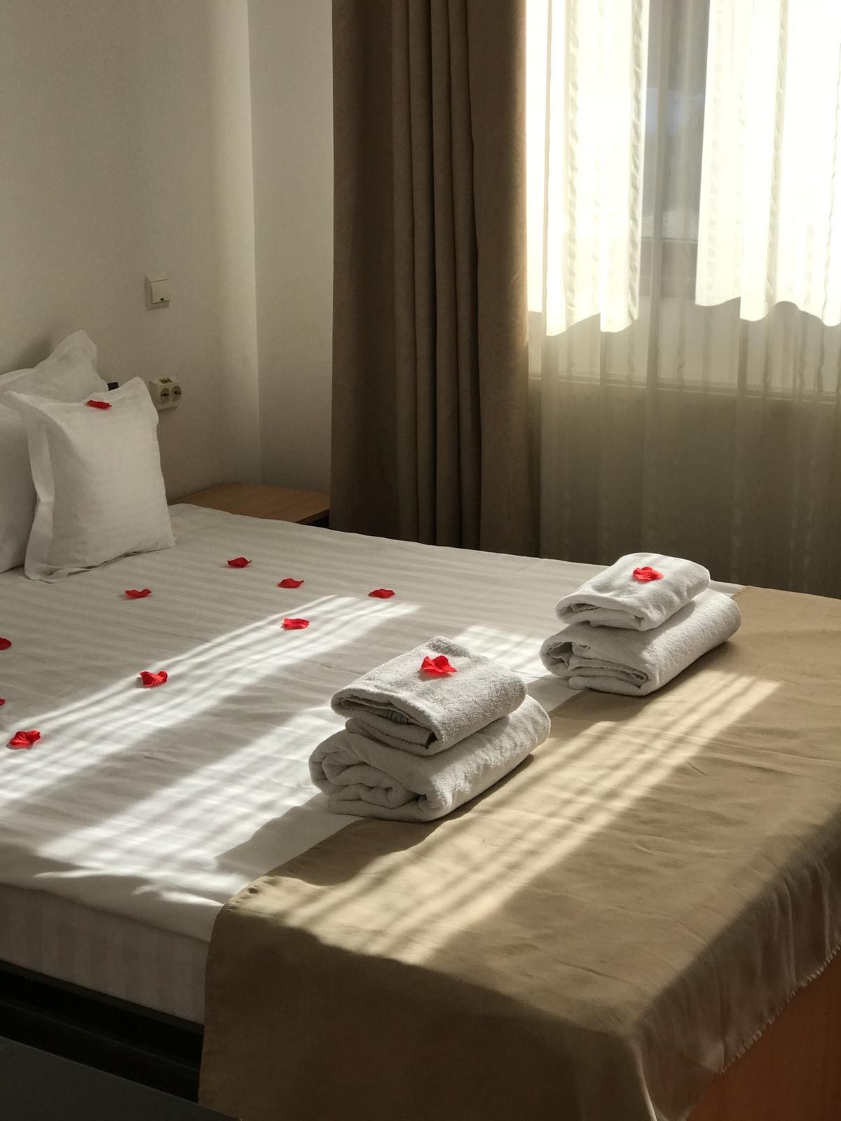 A well-appointed bedroom features a spacious bed adorned with white linens and decorative petals. Neatly folded towels are arranged on the bed. Natural light is filtered through sheer curtains, casting soft shadows across the room.