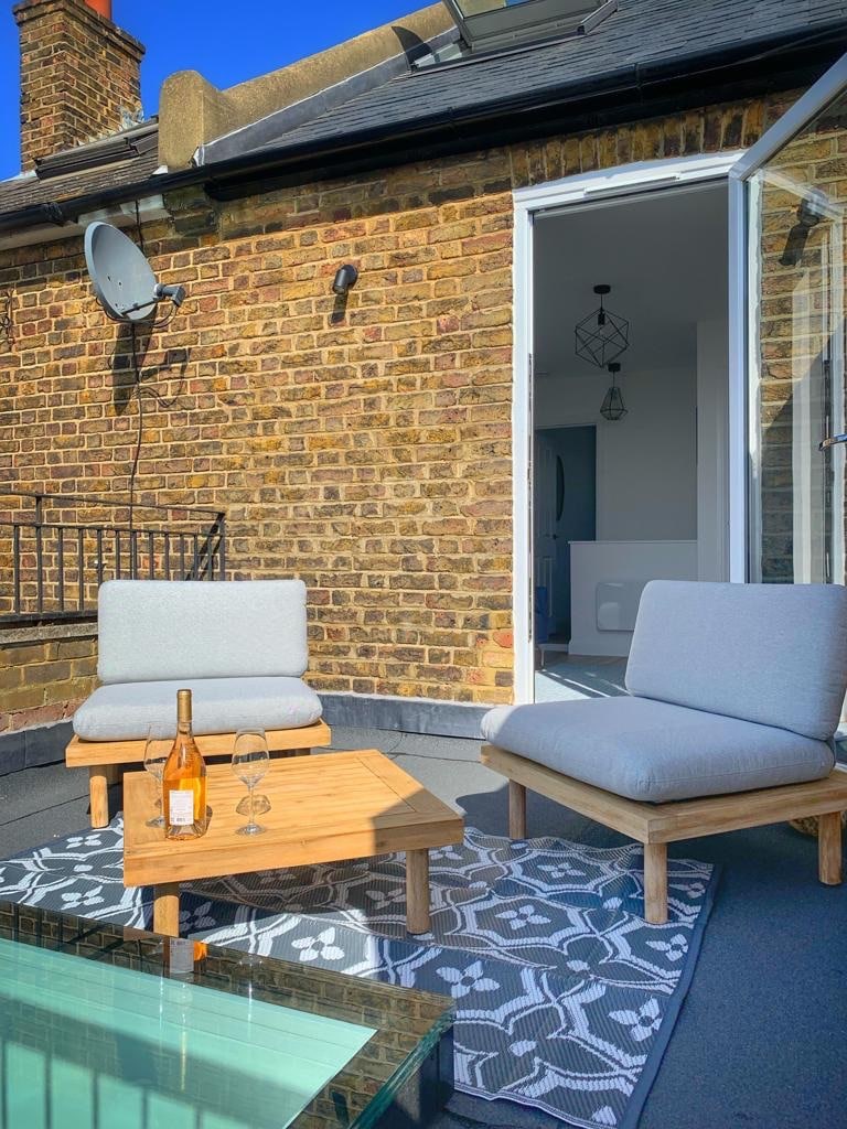 An outdoor balcony features two light grey armchairs positioned around a natural wood coffee table. A bottle of rosé wine and two glasses sit on the table, with a patterned outdoor rug beneath. The view includes brick walls, adding a cozy touch.