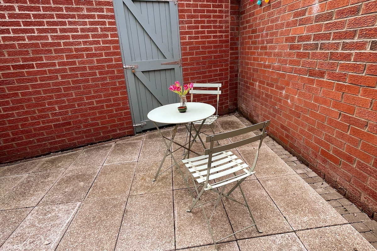 A small outdoor patio area features a round table accompanied by two folding chairs. A potted plant with vibrant flowers sits on the table. The space is bordered by a textured red brick wall and includes a grey garden gate in the background.
