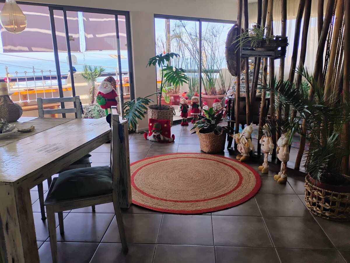 A bright dining area features a wooden table surrounded by chairs, complemented by natural light streaming through large windows. Potted plants and decorative figurines add a touch of greenery and charm. A circular area rug in red outlines the seating space, enhancing the room's layout.