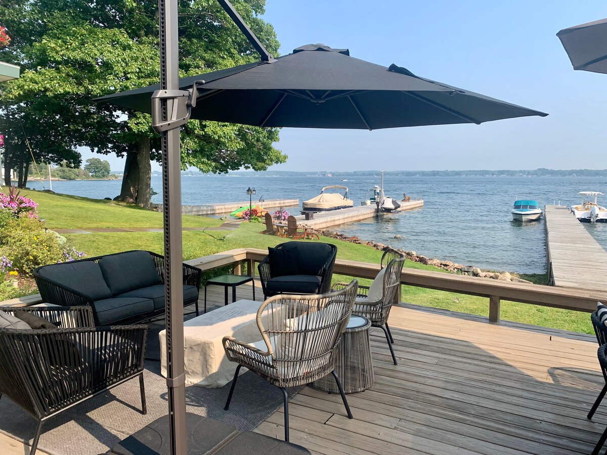 A spacious deck is equipped with comfortable seating arrangements, including a cushioned sofa and woven chairs. An umbrella provides shade over a table. The serene view of the St. Lawrence River channel is framed by greenery and a distant dock for boats.