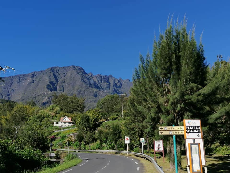 The image displays a scenic road leading towards a mountainous backdrop. A well-maintained house can be seen in the distance, surrounded by lush greenery. Road signs provide directions, and tall trees line the roadside, contributing to the natural charm of the landscape.