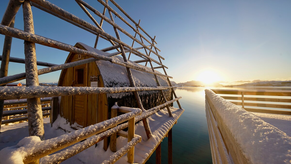 A charming rorbu, partially nestled in snow, is framed by wooden beams and overlooks a serene body of water. The sun rises in the distance, casting a gentle glow on the surrounding landscape. A railing leads towards the structure, enhancing the tranquil setting.