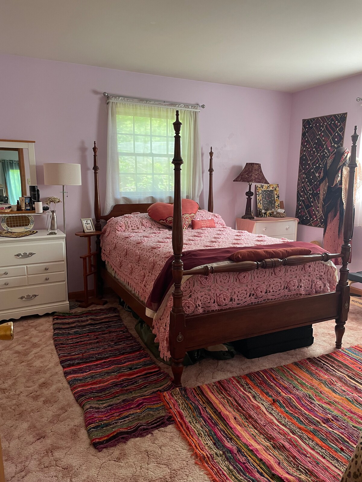 A cozy bedroom features a wooden four-poster bed adorned with a pink lace coverlet and decorative pillows. A window with sheer curtains allows natural light to brighten the space. A colorful woven rug lies on the floor, complementing light purple walls.
