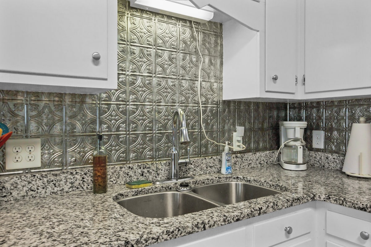 A modern kitchen showcases a polished granite countertop with a double sink. The backsplash is adorned with detailed metallic tiles, reflecting light. A coffee maker and a bottle are placed on the counter, alongside an integrated power outlet.