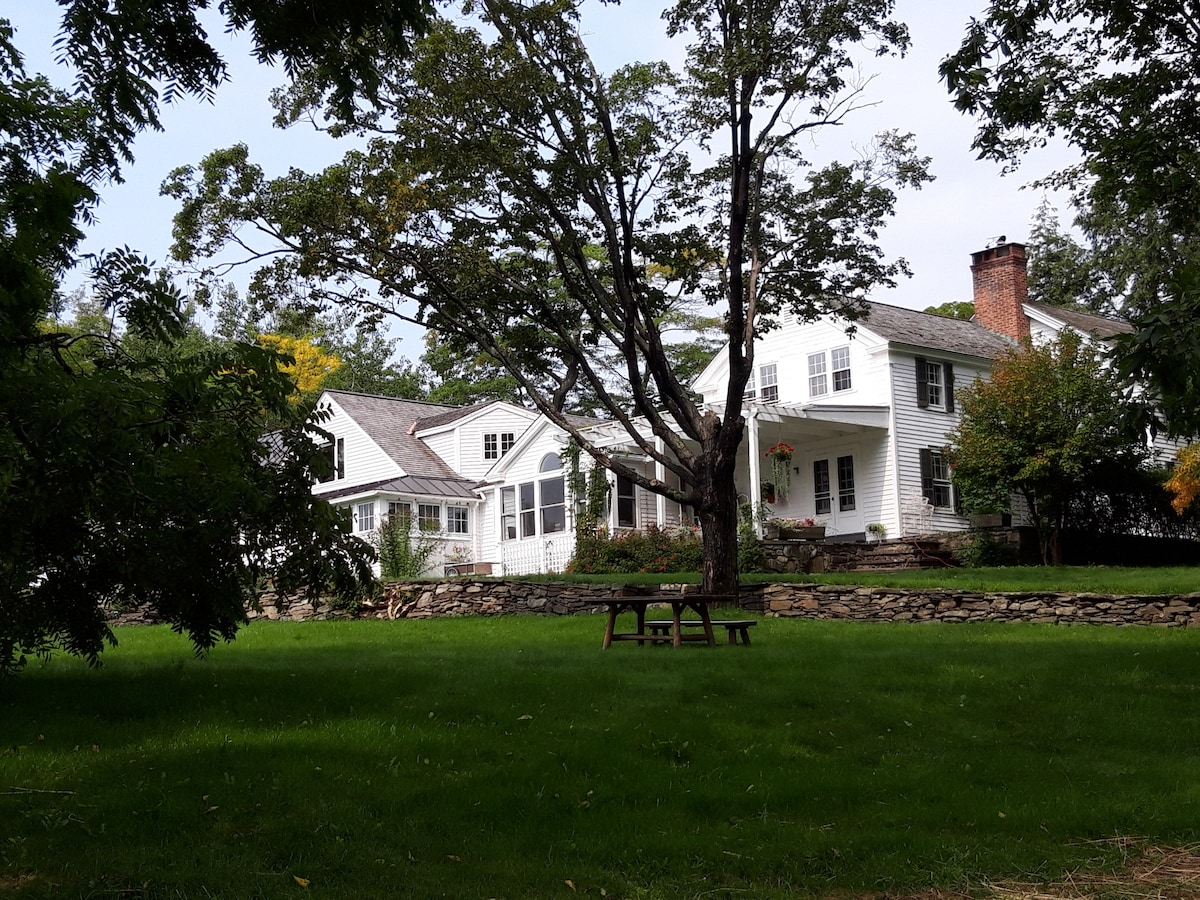 A spacious white house is set among lush green lawns and mature trees, featuring multiple gabled roofs. A large stone wall runs along the property, and a picnic table is placed on the grass, providing a serene outdoor setting.