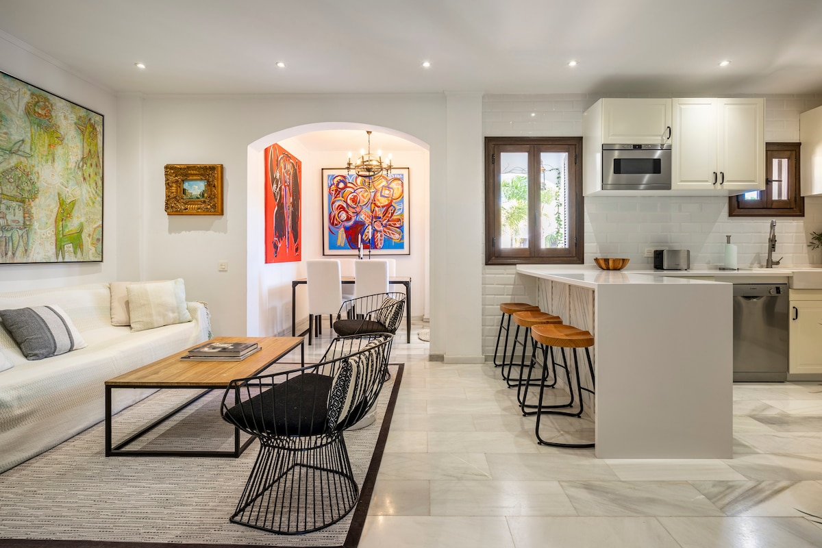 A modern living area displays a mix of contemporary art and minimalist design. A light-colored sofa complements a wooden coffee table placed on a textured rug. High stools are positioned around a kitchen island, while natural light enters through multiple windows, brightening the space.