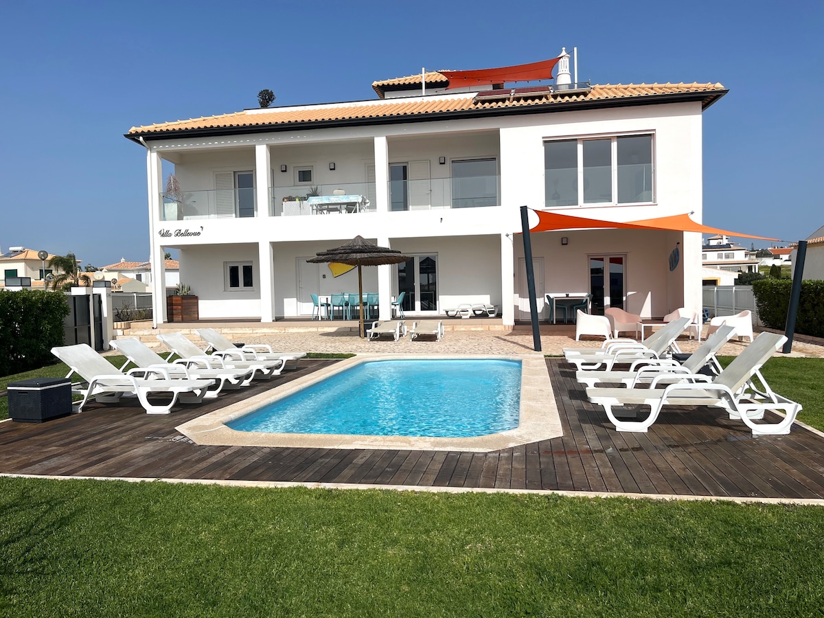 The exterior view showcases a modern two-story house with large glass doors. A rectangular salt-water pool is framed by a wooden deck and surrounded by several lounge chairs. The well-kept garden includes a shaded area, with vibrant colors from the awning enhancing the outdoor space.
