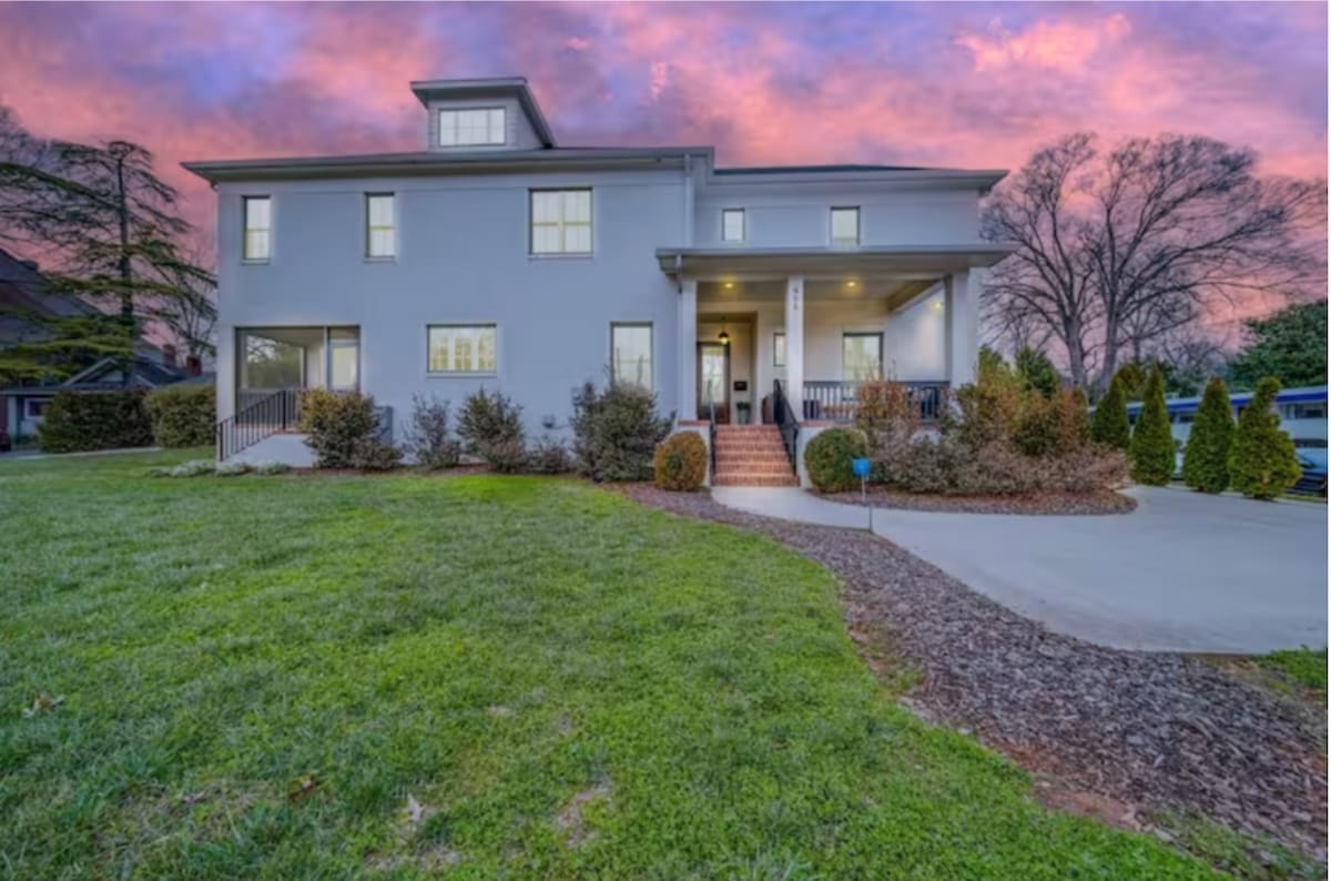 The exterior of the townhome is shown, featuring a light blue facade and multiple windows. A welcoming front entrance can be seen with steps leading up to the door. Lush green grass and landscaped shrubs create an inviting outdoor space, with a colorful sunset sky in the background.