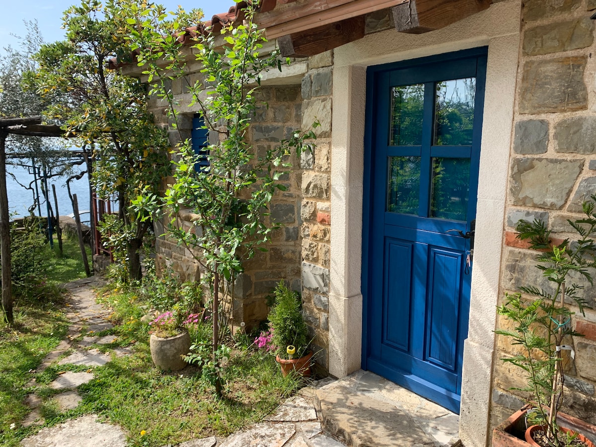 A vibrant blue door frames the entrance of the small summer house, surrounded by stone walls and lush greenery. A path made of stone leads to the door, with colorful flowers and plants positioned along the side, enhancing the natural setting by the bay.