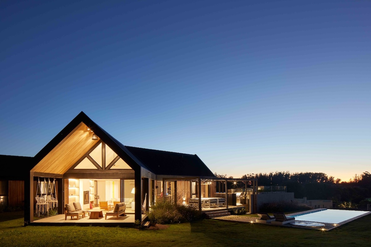 The exterior of a modern home is depicted at dusk, featuring a gabled roof and expansive windows that allow warm interior light to shine through. A pool is visible in the foreground, reflecting the twilight sky, while comfortable outdoor seating is arranged nearby.