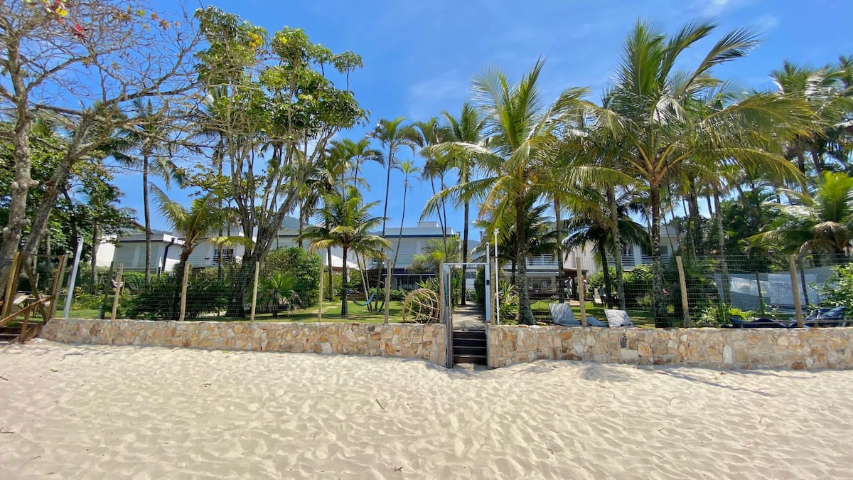A serene beachfront property is framed by palm trees and lush greenery. A sandy pathway leads from the beach to the house, providing direct access. The scenic outdoor area includes a stone wall and is set against a clear blue sky.