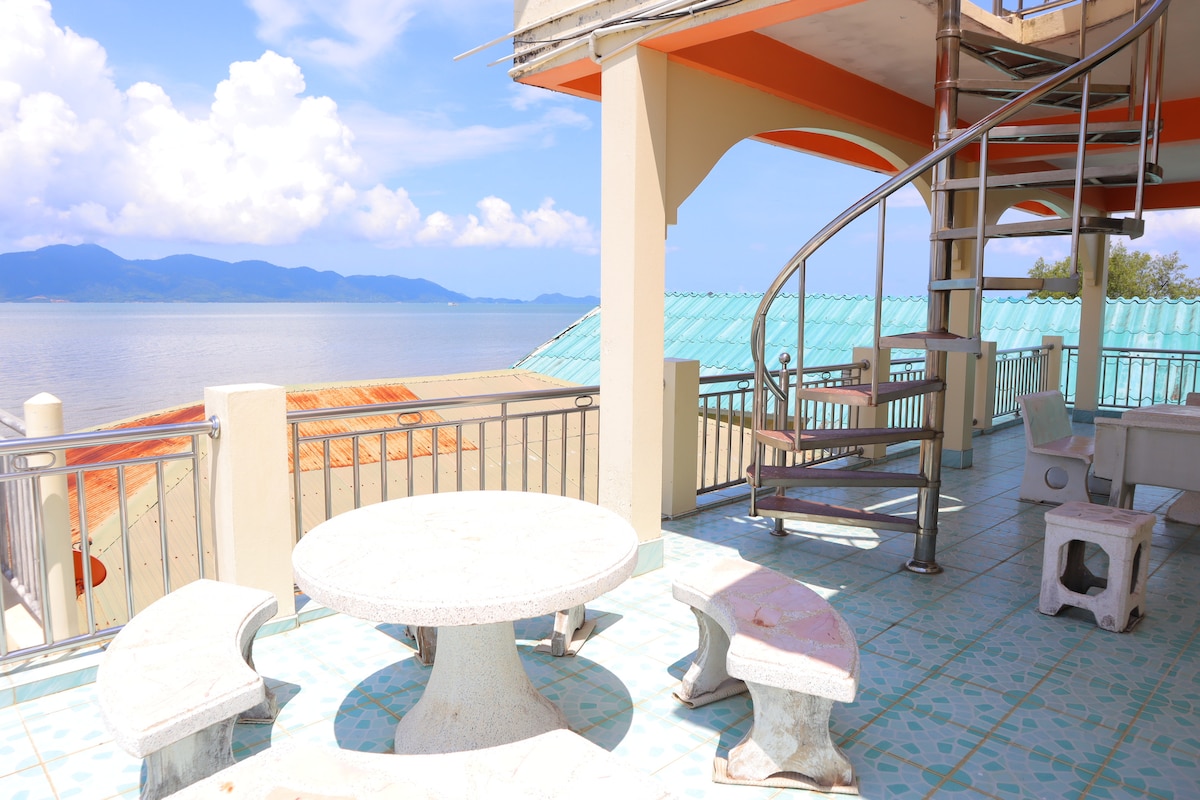 An outdoor deck area features a round table with matching stone benches, providing a place for seating and relaxation. A spiral staircase leads to an upper level. Scenic views of the water and mountains are visible in the distance, embraced by clear blue skies.