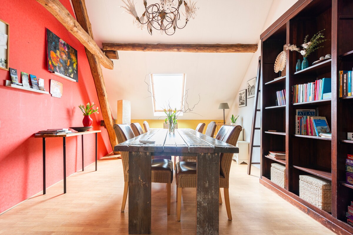 A spacious dining area features a large wooden table surrounded by several chairs. Bright natural light streams through an overhead window, illuminating the room's vibrant red and neutral-colored walls. A bookshelf filled with various items stands against one wall, complemented by decorative elements.