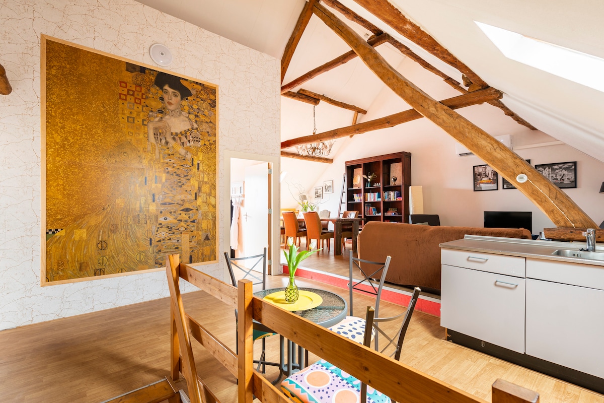 A spacious open-plan area is highlighted by a large artwork on one wall and airy, wooden beams overhead. The kitchen features modern appliances, while a dining area with a large table invites communal gatherings. A comfortable living space is also visible.