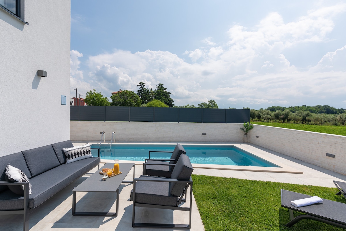An outdoor seating area features modern grey lounge furniture arranged around a low table. A private swimming pool is visible in the background, surrounded by landscaped grass and a privacy fence. The sky is partly cloudy, enhancing the serene atmosphere.