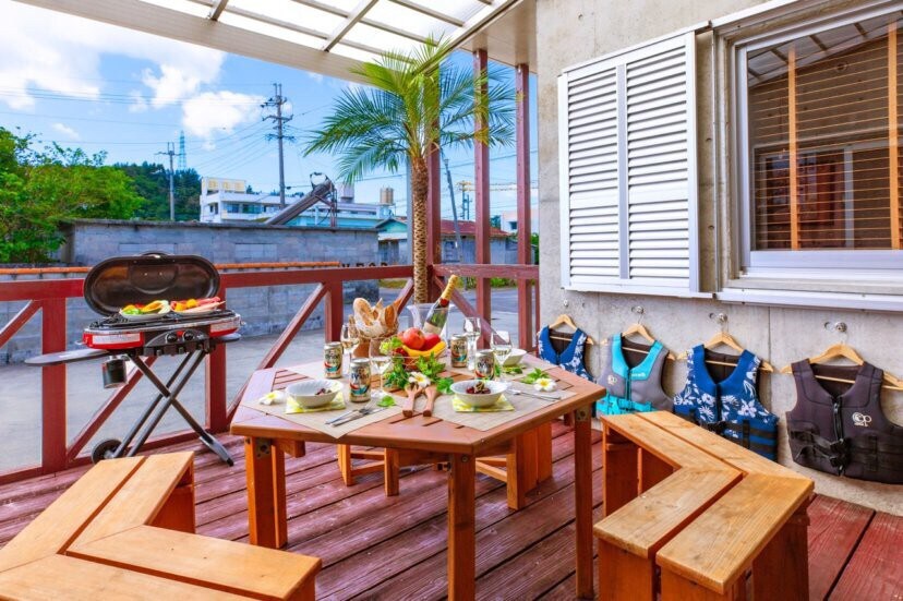 An outdoor dining area features a wooden table surrounded by benches, set for a meal with plates, glasses, and decorative fruit. A grill stands nearby. Sunlight filters through a shaded roof, while palm trees and colorful life jackets add a relaxed atmosphere.