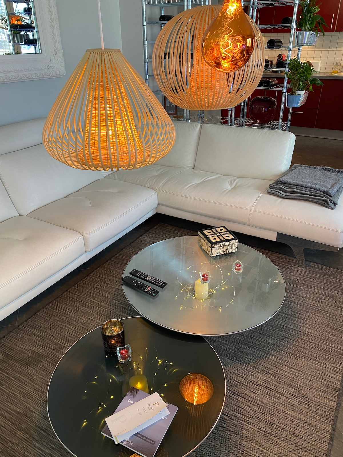 A spacious seating area features a white, curved sectional sofa positioned around two round glass coffee tables. Unique woven pendant lights cast a warm glow above the setting. A stack of folded blankets and a remote control are placed casually on the sofa.
