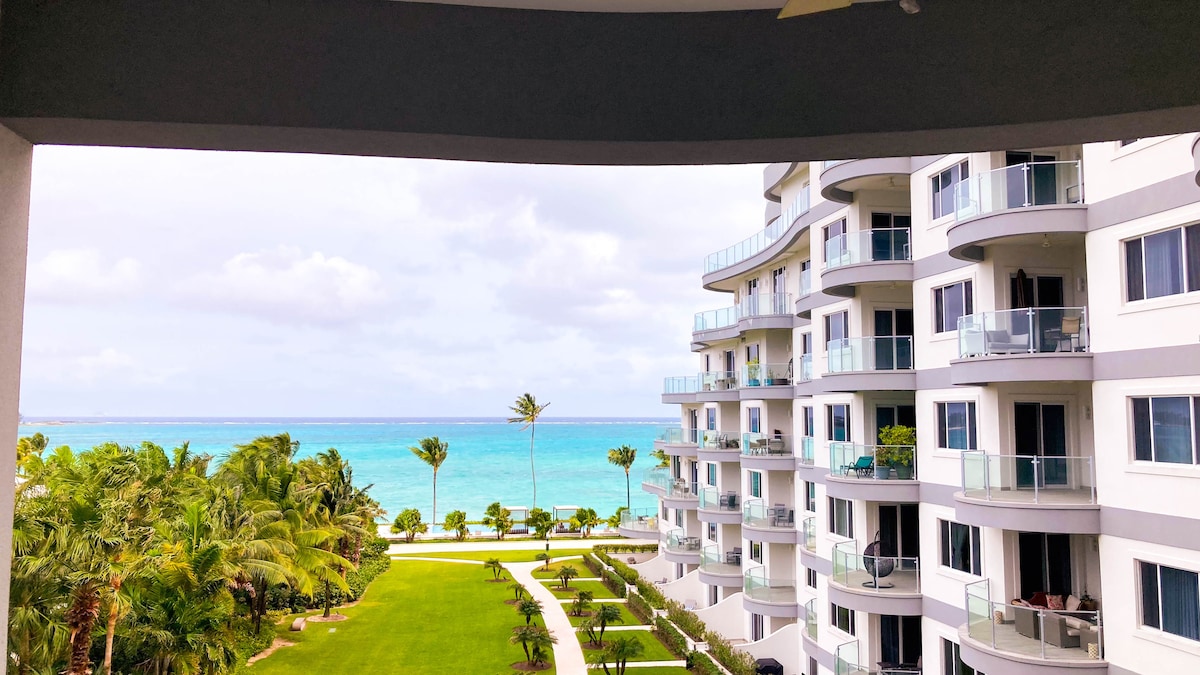 A balcony view reveals a serene ocean stretching towards the horizon, complemented by lush green palm trees and meticulously maintained gardens. The modern condominium structure curves elegantly, featuring glass railings that enhance the coastal atmosphere.
