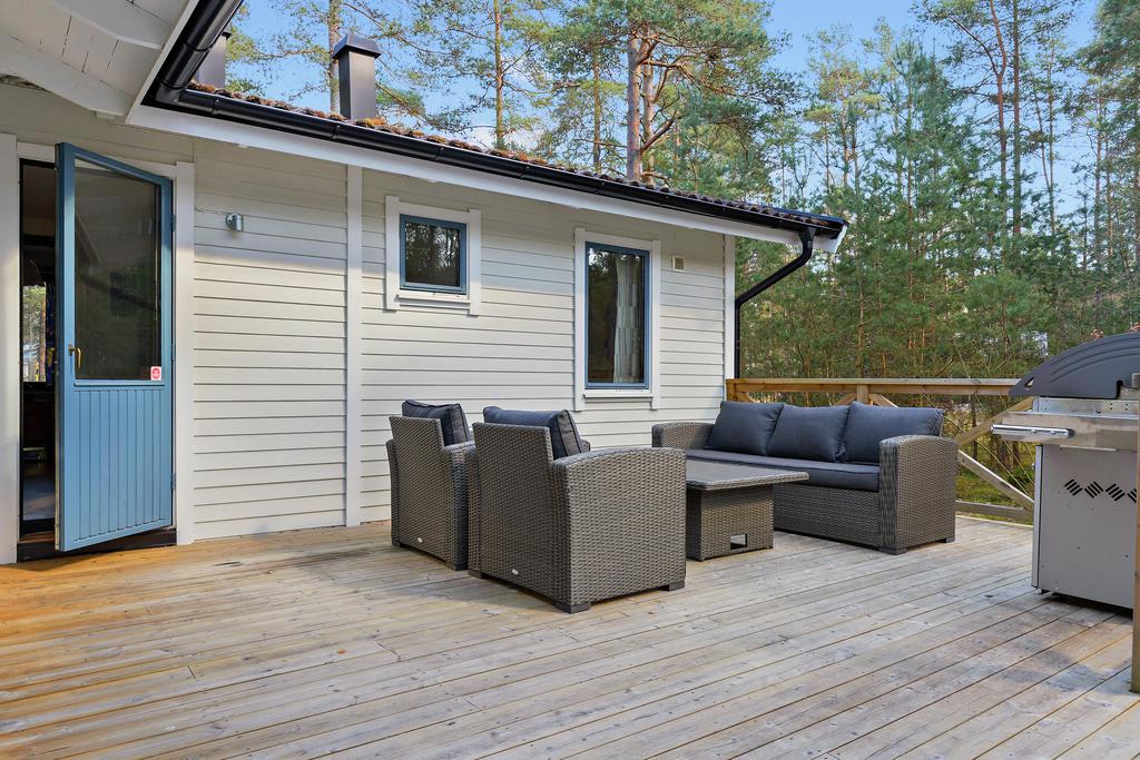 An outdoor seating area features a comfortable, woven sofa set arranged around a low table, positioned on a wooden deck. Two windows are present on the exterior wall, allowing natural light into the space. A grill is situated in the corner of the deck.