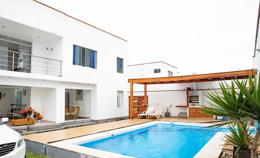 The outdoor area features a rectangular swimming pool surrounded by a wooden deck. A pergola provides shade over a grilling area. White walls of the building reflect natural light, enhancing the spacious appearance. Colorful plants are situated near the pool, adding a touch of greenery.