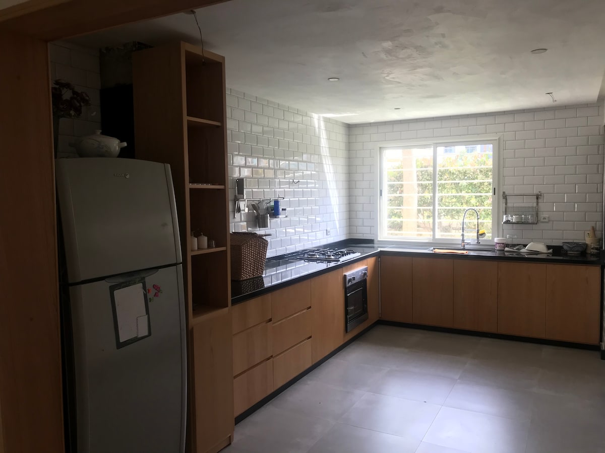 A modern kitchen features sleek cabinetry and a spacious countertop. A stainless steel refrigerator and an oven are visible, alongside a gas stove. Natural light enters through a window, illuminating the white tiled walls and providing a clean and bright cooking environment.