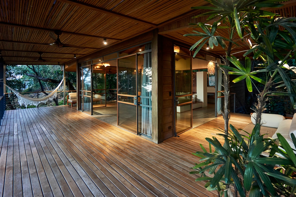 A spacious outdoor terrace features wooden flooring and large glass doors that open to an interior space. Lush greenery surrounds the area, with a hammock suspended between two posts. Comfortable seating options invite relaxation, enhancing the connection with nature.